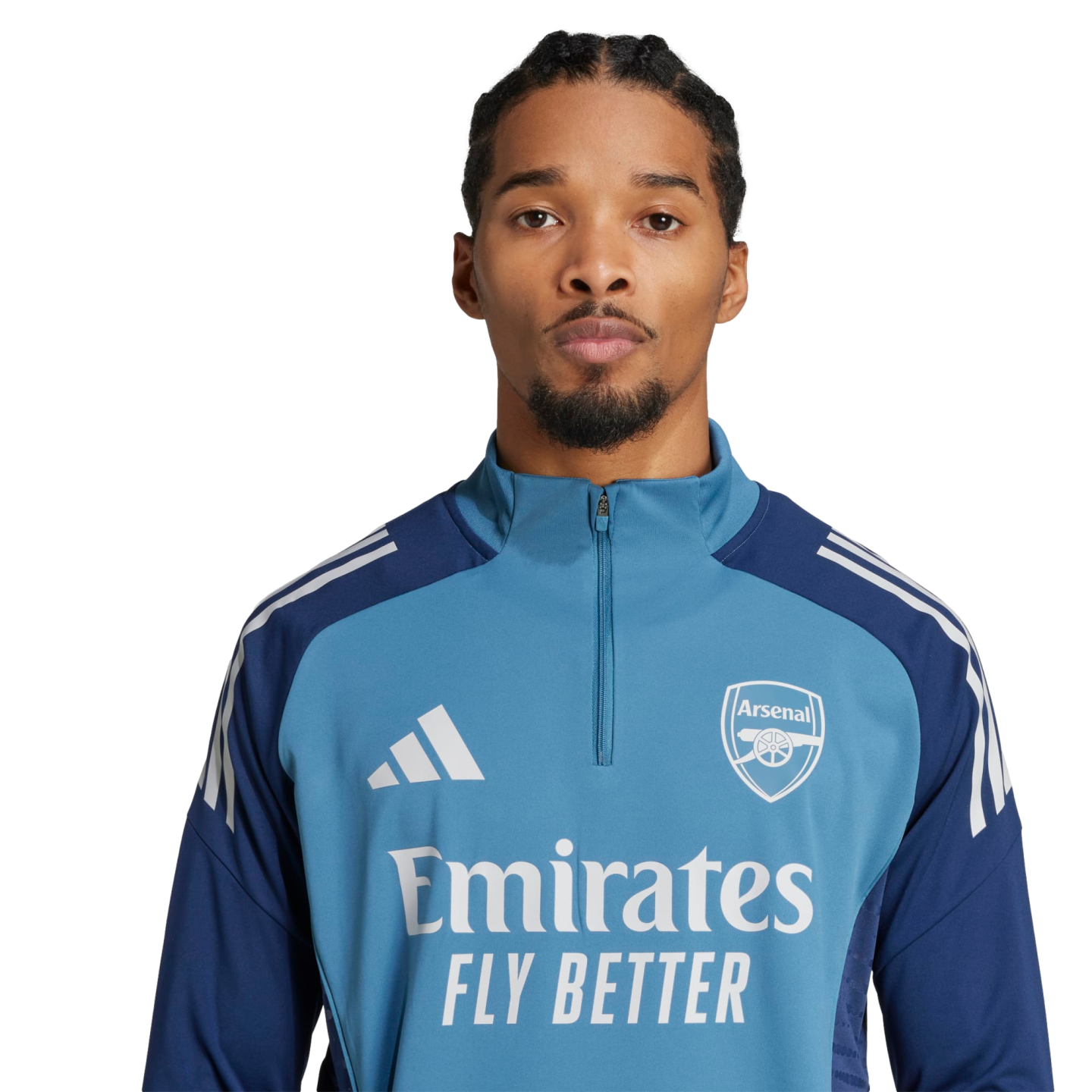 Headshot of a man with braided hair and a goatee wearing a blue Arsenal training zip-up top with the Emirates sponsor logo, set against a solid black background.
