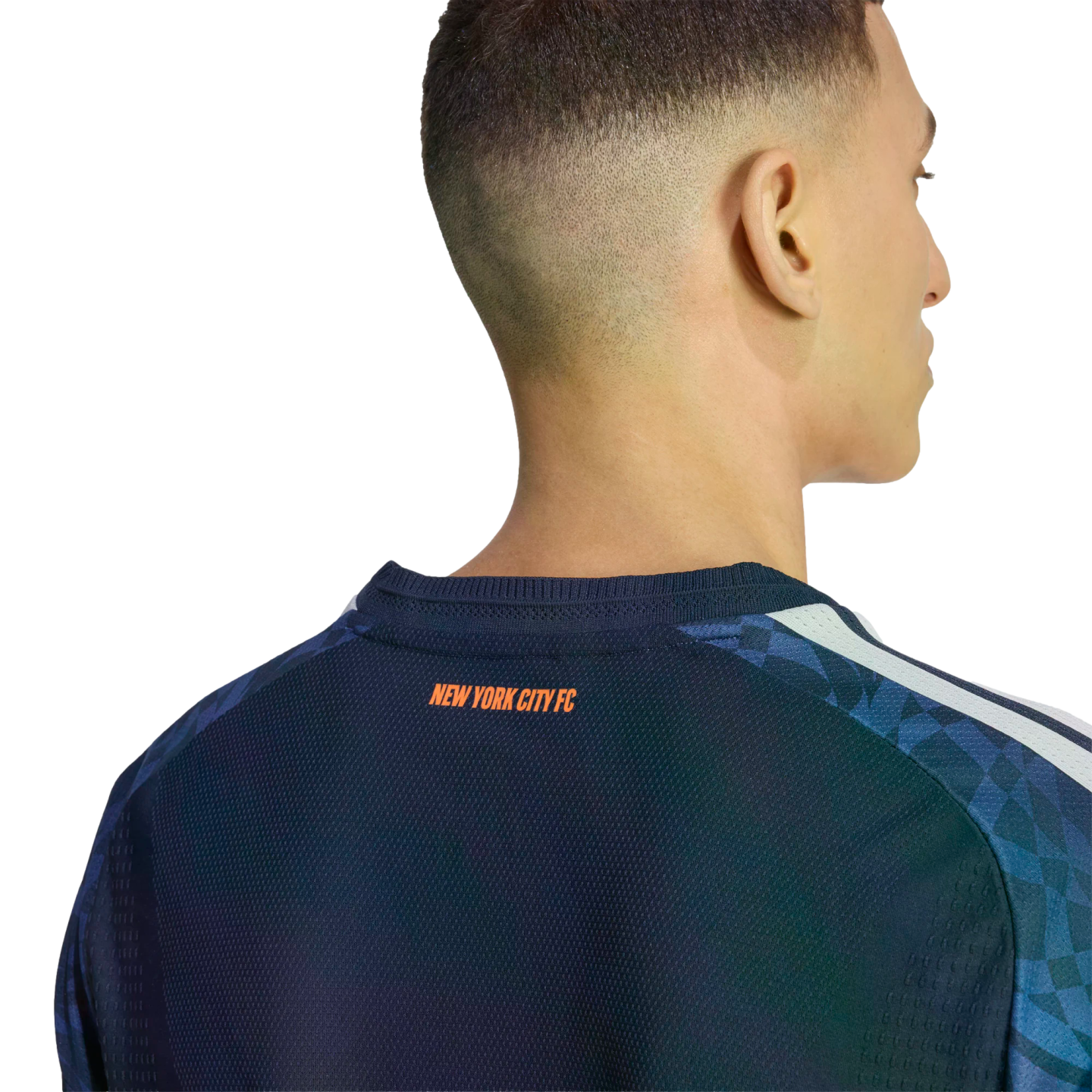 Back view of a man wearing a navy New York City FC soccer jersey with orange text below the collar. The shoulders have a blue geometric pattern and white stripes.
