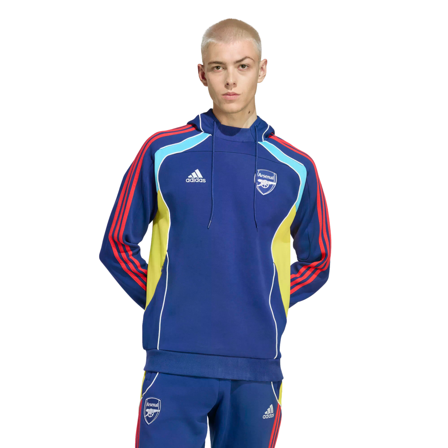 A man with buzzed blonde hair wearing a blue Adidas Arsenal FC tracksuit with red stripes and yellow/light blue accents, posed against a black background.