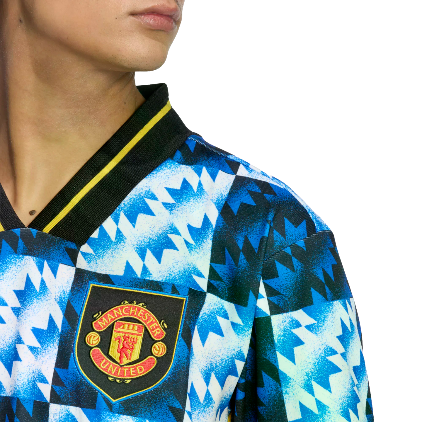 Close-up of a Manchester United jersey with a blue and white jagged geometric pattern and the club crest, featuring a black collar with yellow trim.