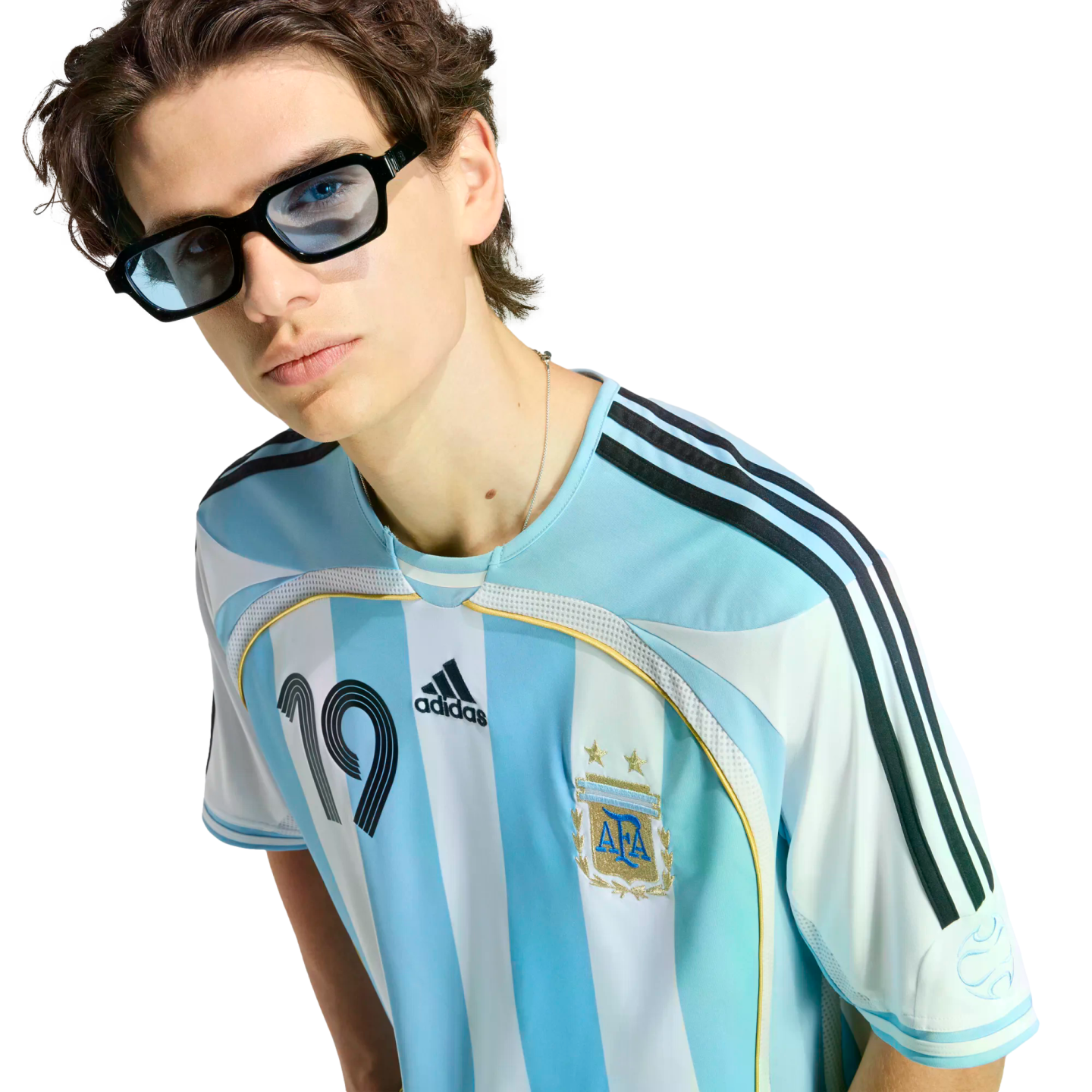 A young man with wavy hair and blue-tinted glasses wears an Argentina national team jersey featuring the number 19 against a solid black background.