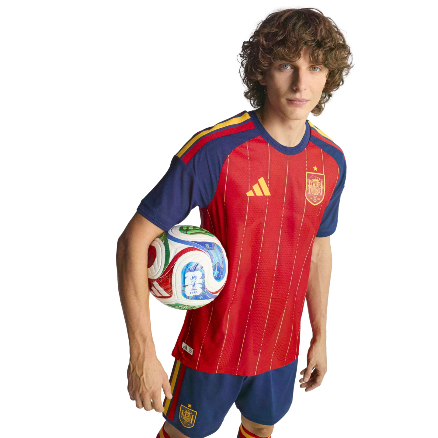A young man in a red and blue Spain national football kit holds a soccer ball under his arm against a black background.