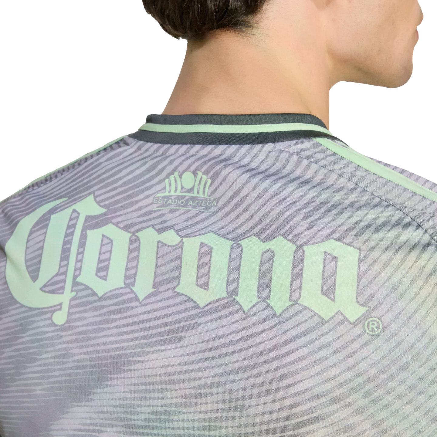 A close-up of the back of a person wearing a light green and grey patterned sports jersey with an Estadio Azteca emblem and a large Corona logo.