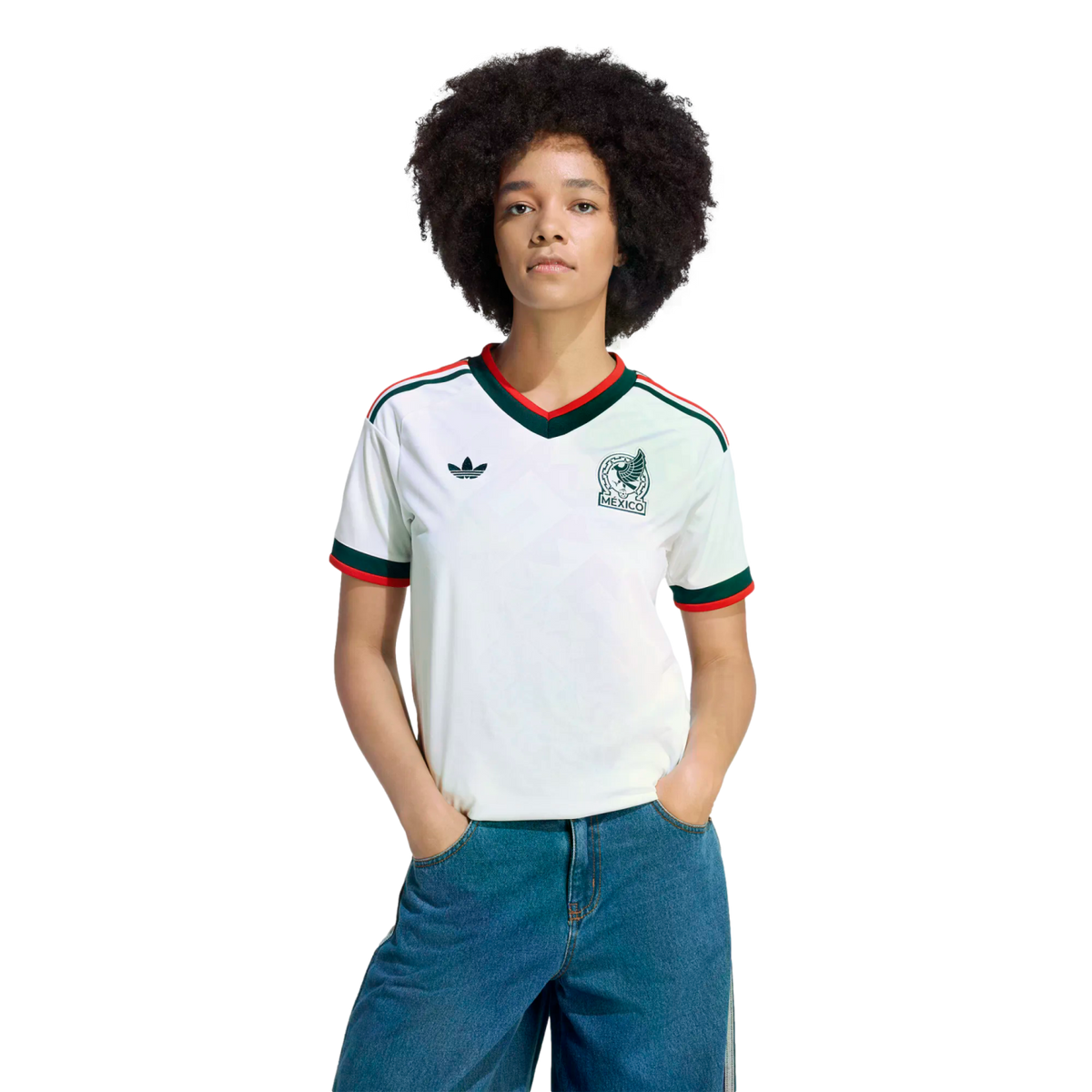 A person with dark curly hair wears a white Mexico Adidas soccer jersey with green and red trim and blue jeans against a solid black background.
