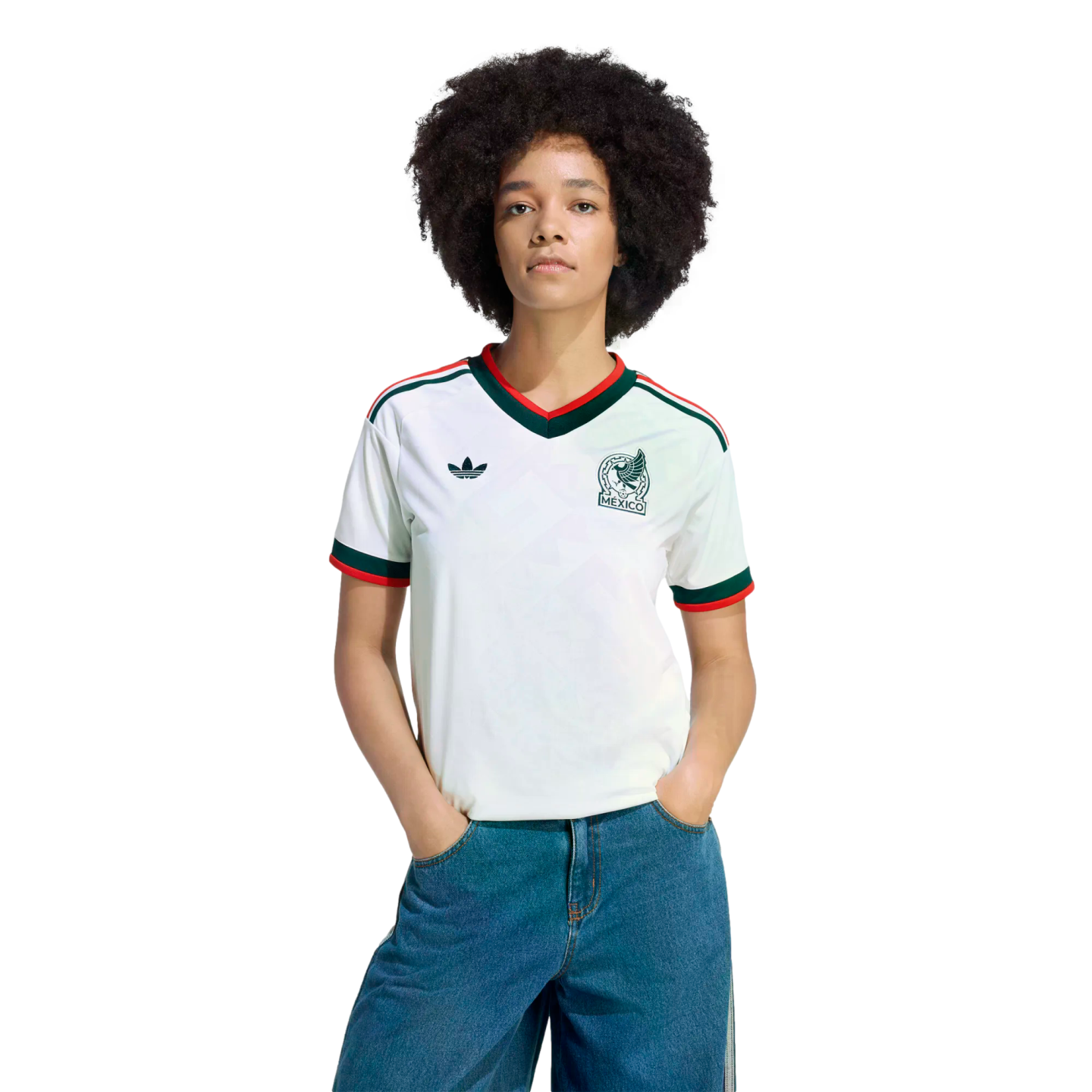 A person with dark curly hair wears a white Mexico Adidas soccer jersey with green and red trim and blue jeans against a solid black background.