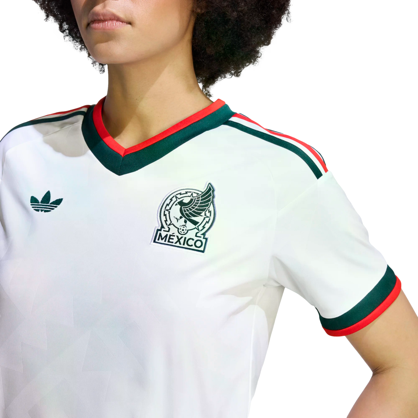 A close-up shot of a person from the chest up, wearing a white Mexico national team soccer jersey by Adidas. The jersey features a green and red V-neck collar, green and red stripes on the shoulders, and matching cuffs on the short sleeves. The Mexico nat