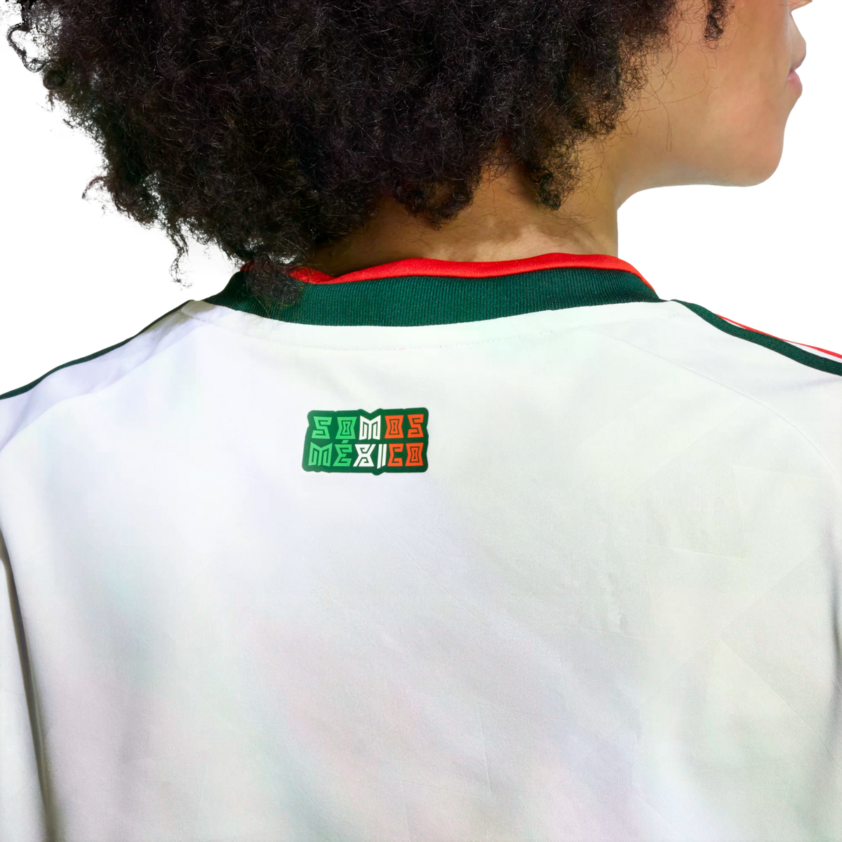 A person with dark curly hair from behind, wearing a white Mexico national team jersey. On the upper back, just below the green and red collar, is a small rectangular logo that reads "SOMOS MÉXICO" in the colors of the Mexican flag.