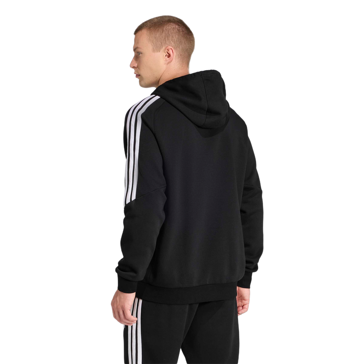 Rear view of a man wearing a black Adidas tracksuit with three white stripes on the sleeves and legs, looking to the side against a plain light grey background.