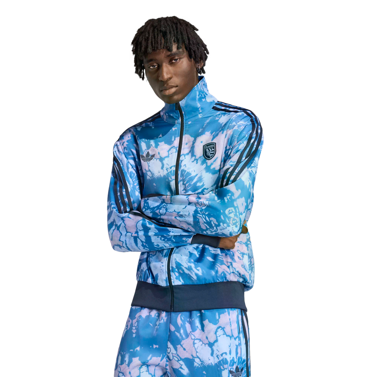A person with dreadlocks poses in a blue tie-dye Adidas tracksuit featuring a "QUAKES" crest, set against a solid black background.