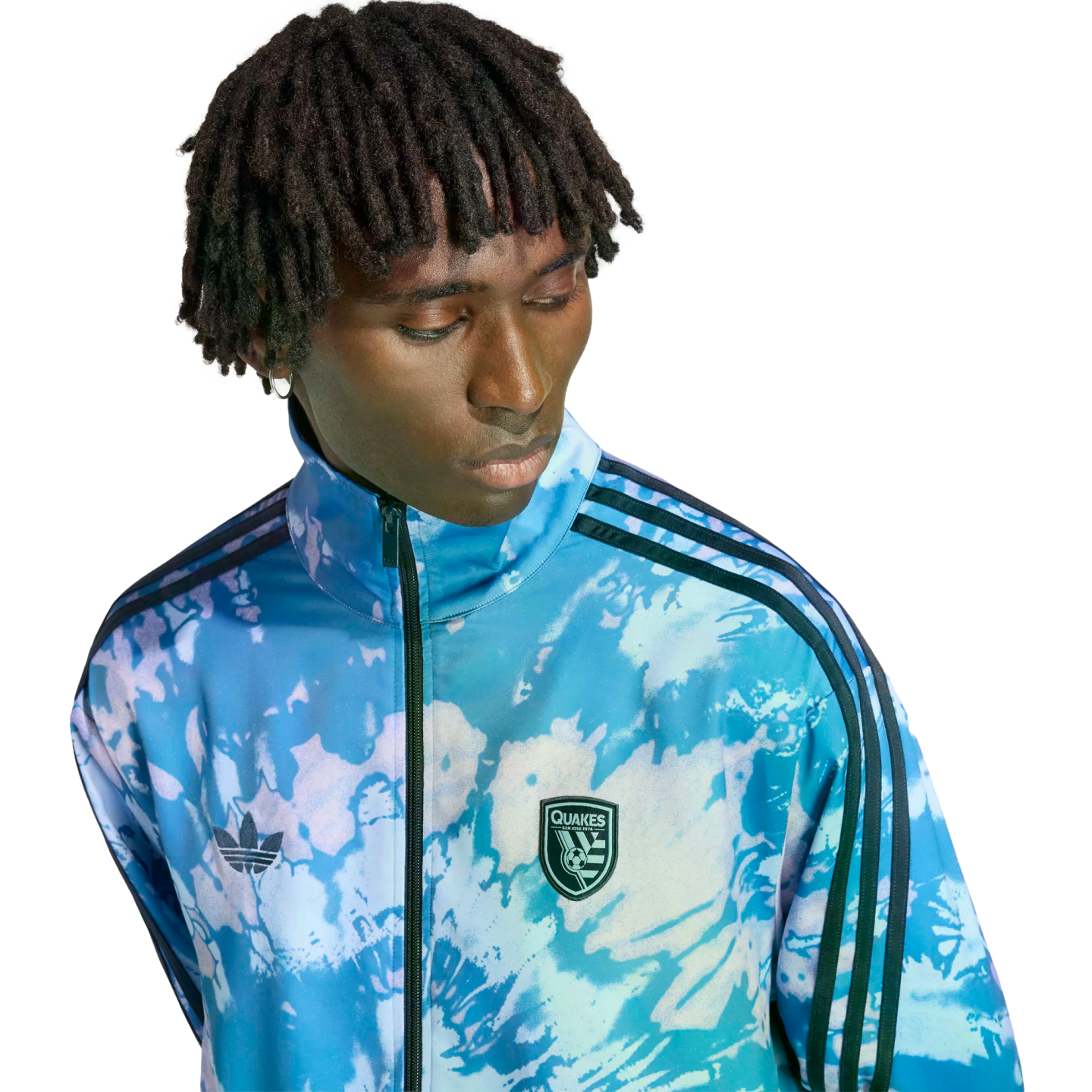 A person with short dreadlocks wearing a blue and white tie-dye patterned Adidas track jacket with a San Jose Earthquakes logo.