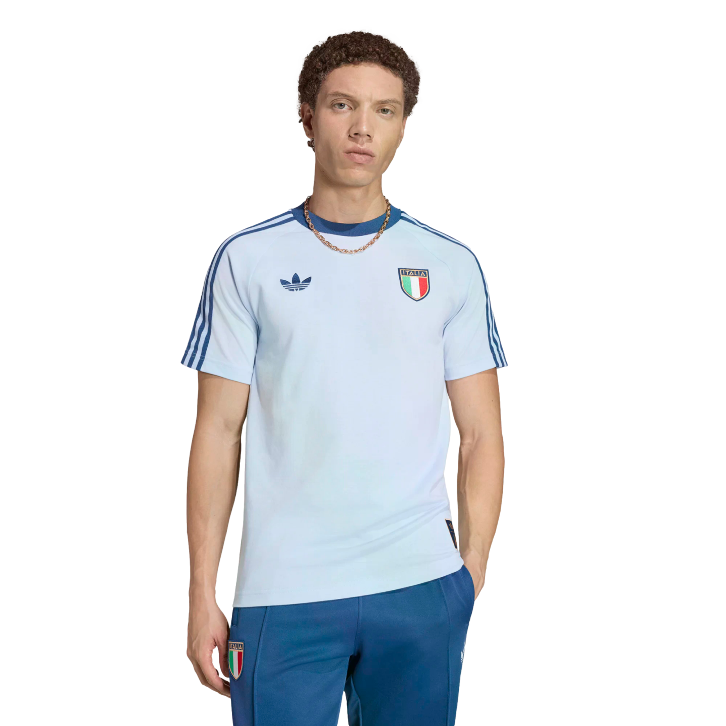 A young man with curly brown hair wearing a light blue Adidas Italy national team t-shirt and dark blue track pants, posed against a solid black background.