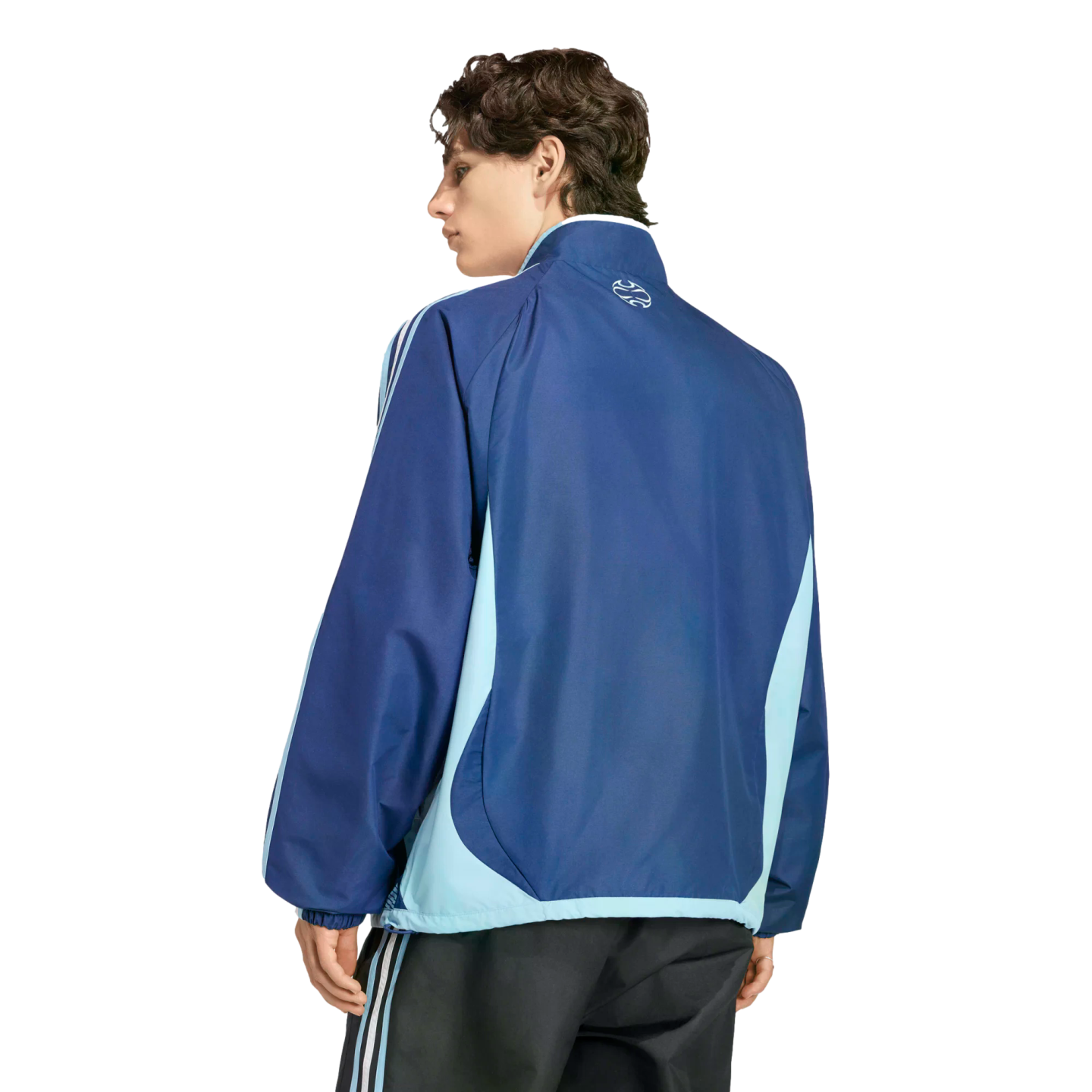 Back view of a man wearing a navy and light blue color-blocked track jacket and dark striped pants, looking over his shoulder against a black background.