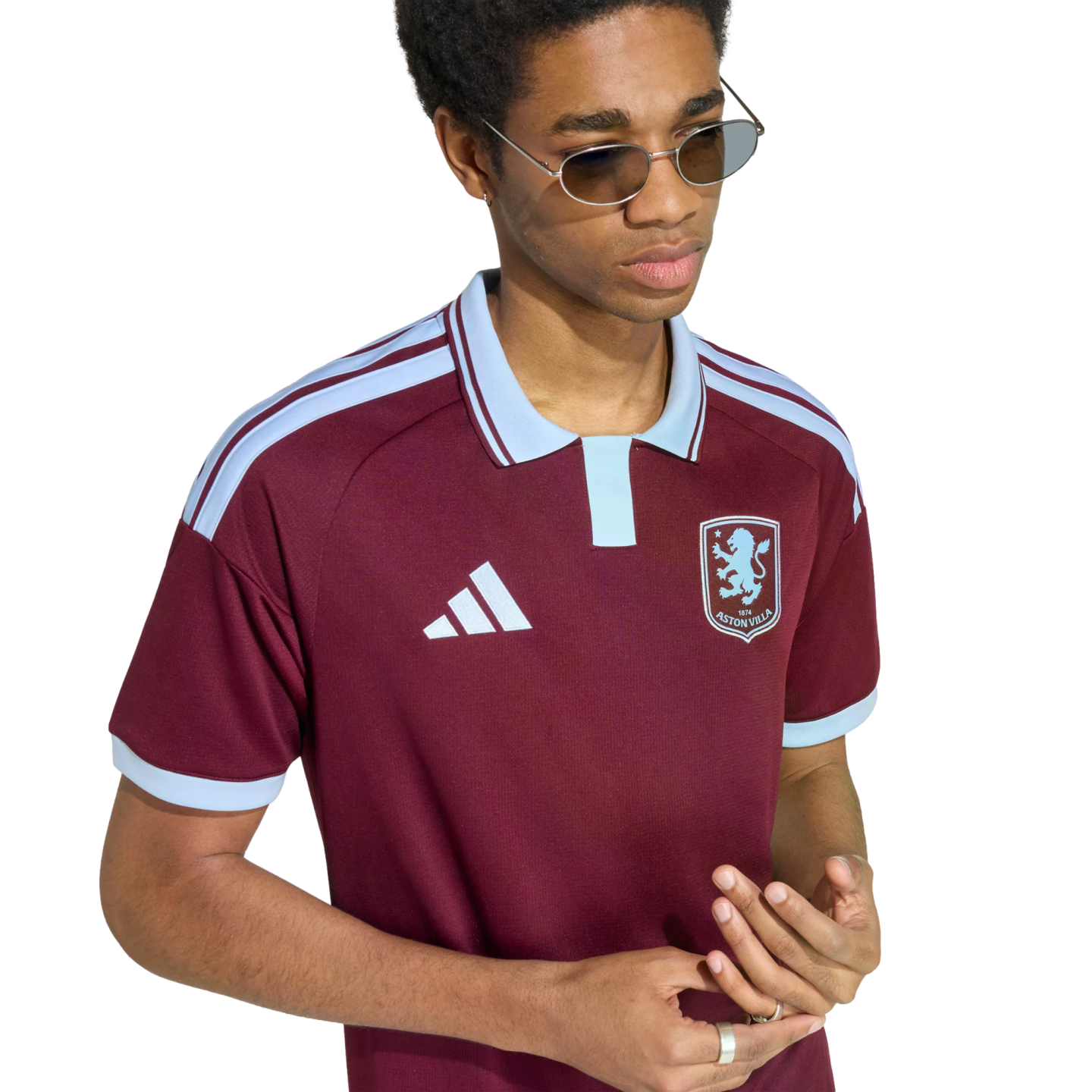 A person with sunglasses and rings wears a maroon and light blue Adidas Aston Villa polo shirt against a plain white background.