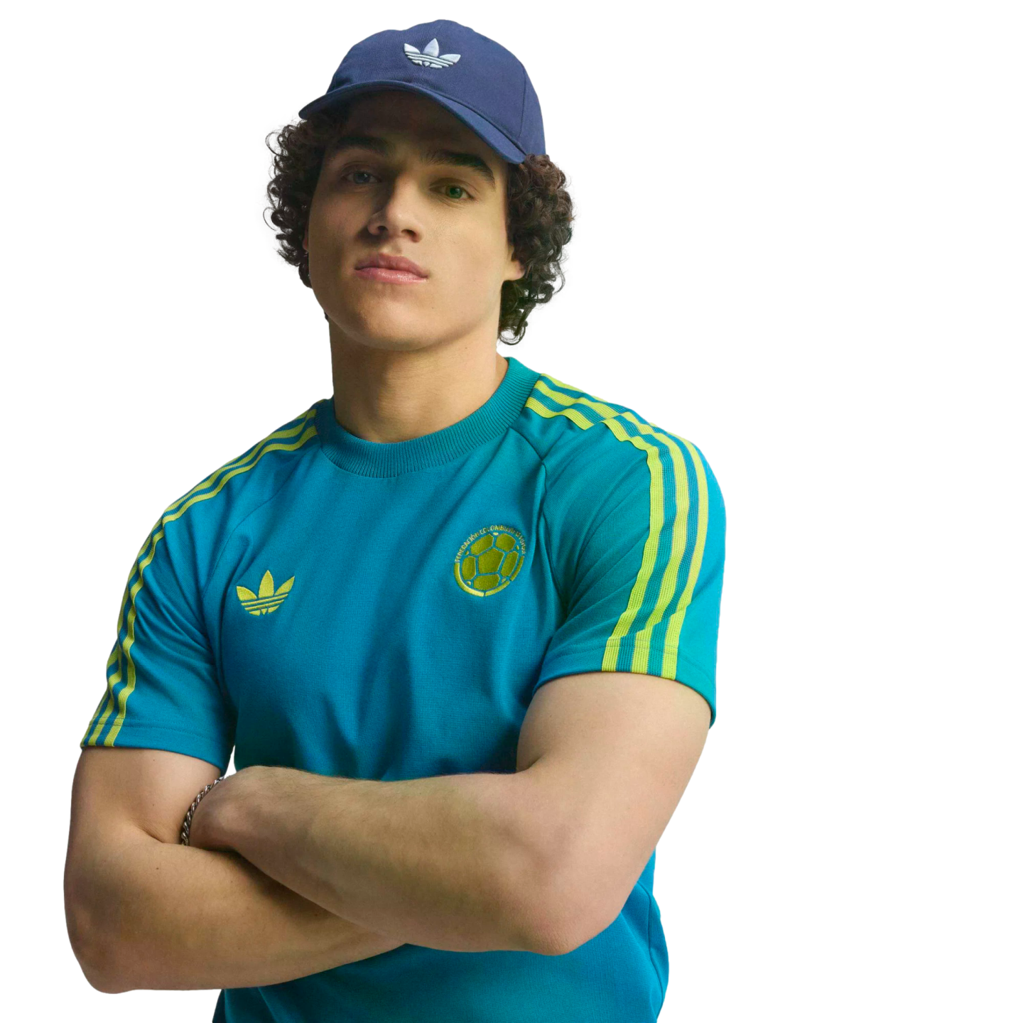 A man in a blue Adidas cap and teal soccer t-shirt with yellow stripes, posing with his arms crossed against a black background.