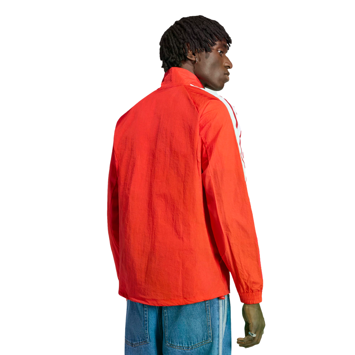 Rear view of a man with dreadlocks wearing a bright orange zip-up jacket and blue jeans against a black background.