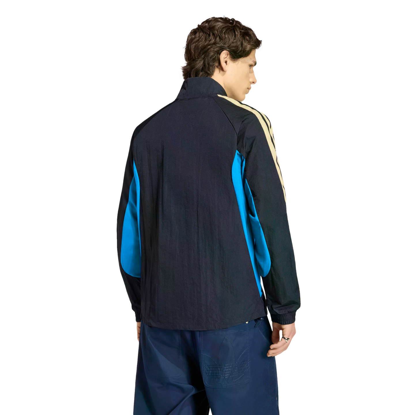 Rear view of a person in a black track jacket with blue side panels and cream stripes, wearing navy blue pants, against a black background.