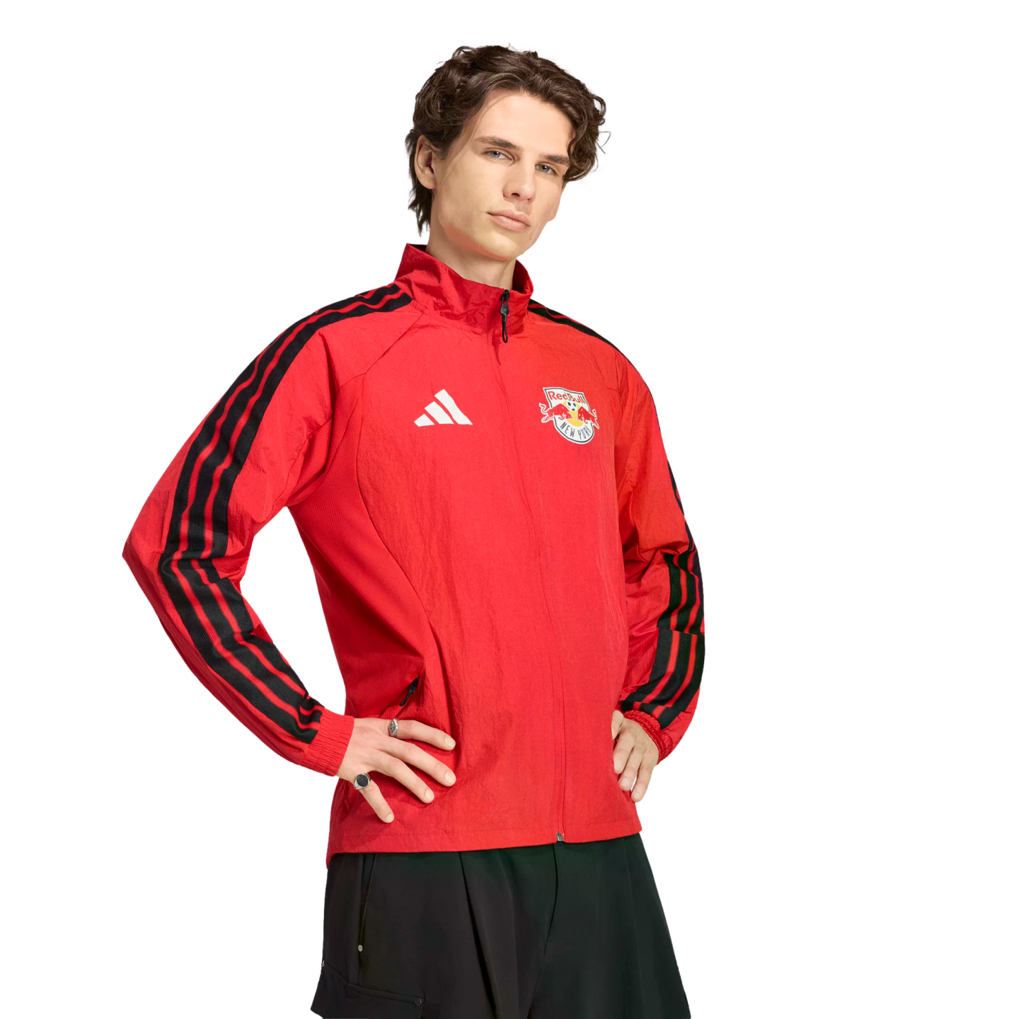 A young man wearing a red New York Red Bulls Adidas track jacket with black stripes, posing with hands on hips against a black background.