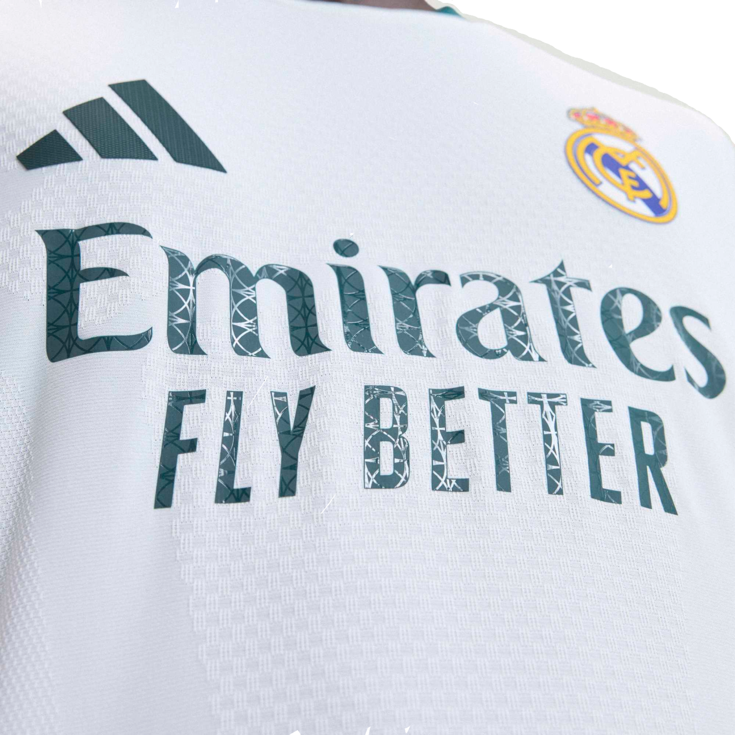 Close-up of a white Real Madrid soccer jersey featuring the Adidas logo, club crest, and "Emirates FLY BETTER" sponsor text with a graphic pattern inside the letters.