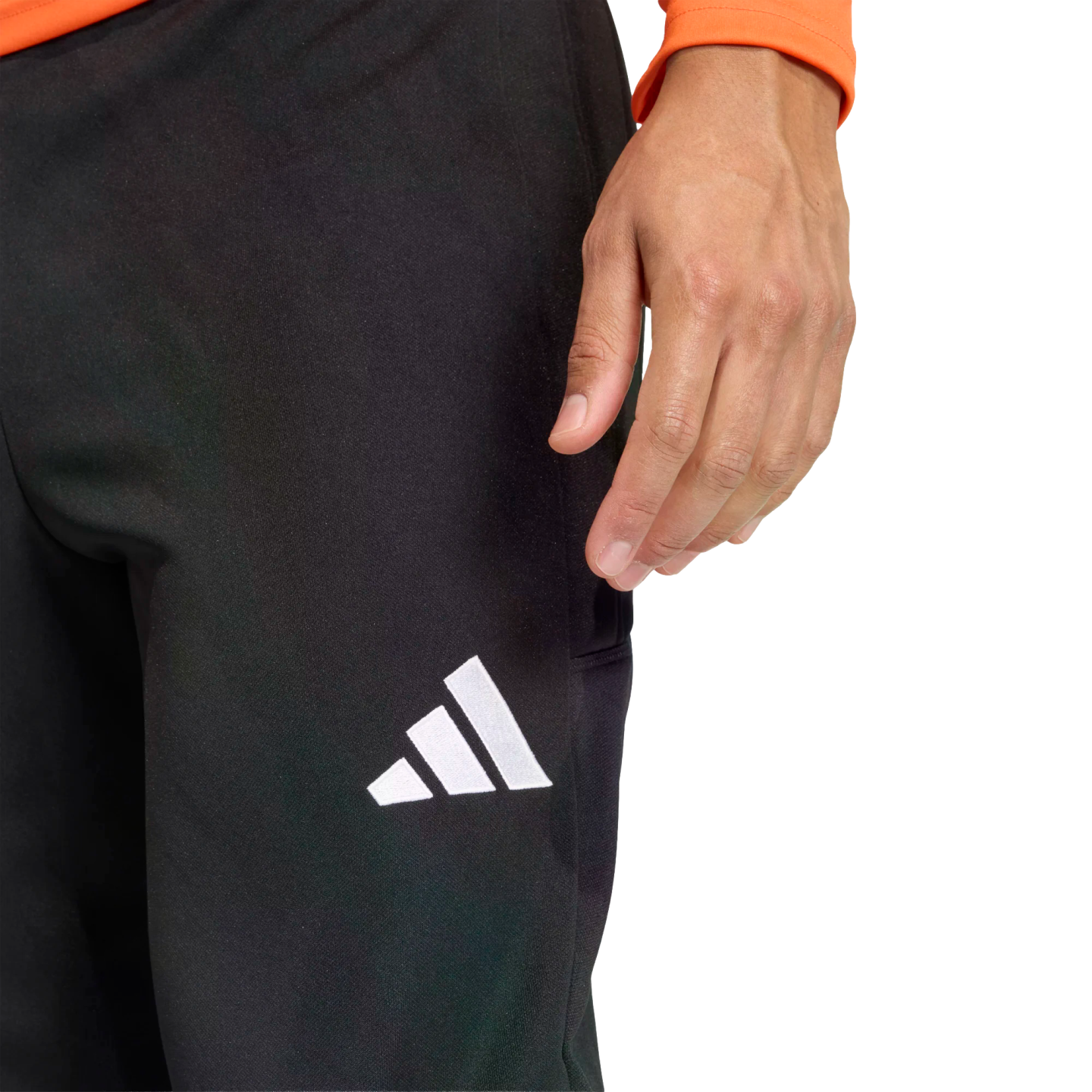 Close-up of a person wearing black Adidas athletic pants with a white logo on the thigh. Their hand rests near their hip, and an orange sleeve is visible against a black background.