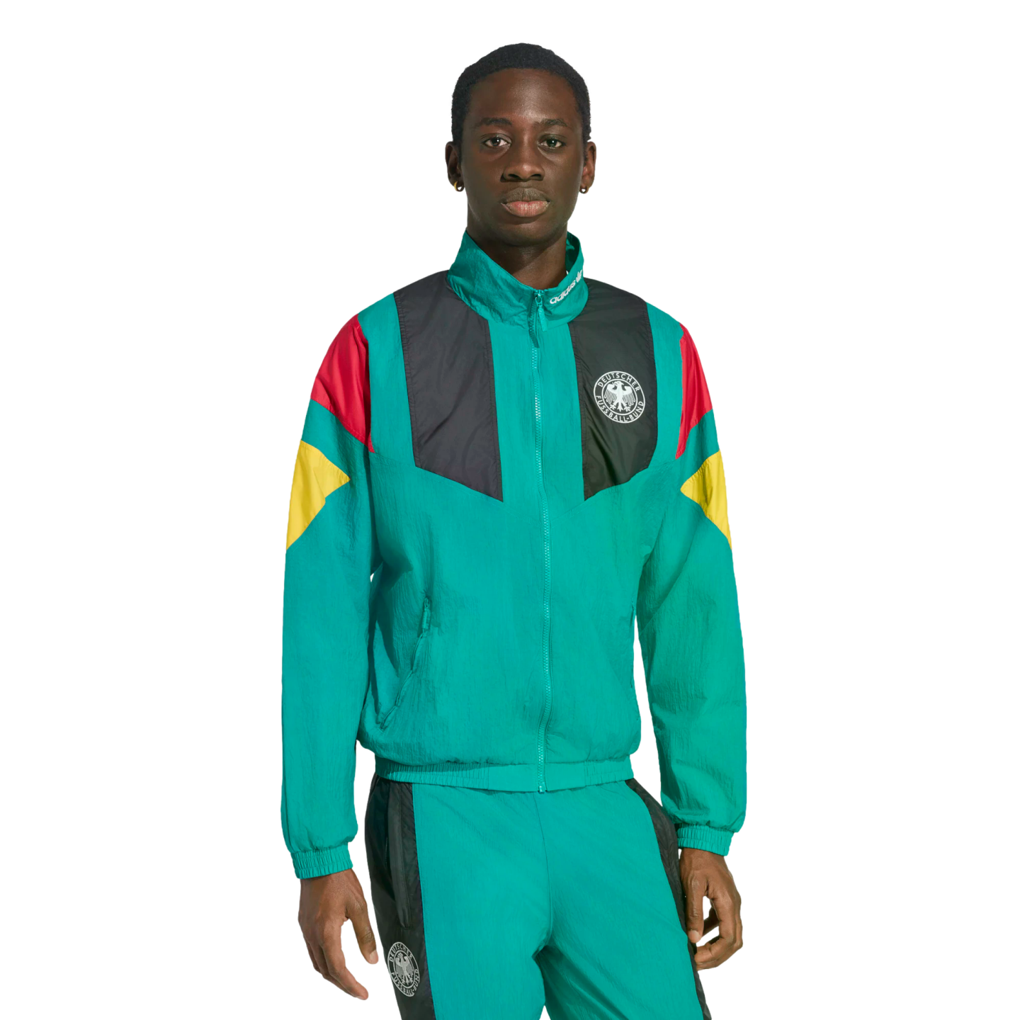 A Black man wears a teal tracksuit with German national team colors and crest on the jacket against a white background.