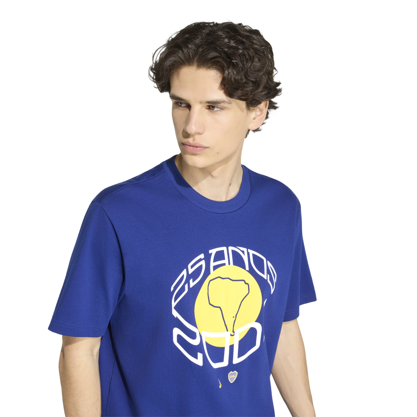 A young man with dark wavy hair wears a blue t-shirt featuring a large graphic with a yellow circle, a map of South America, and the text "25 ANOS 2001" against a white background.