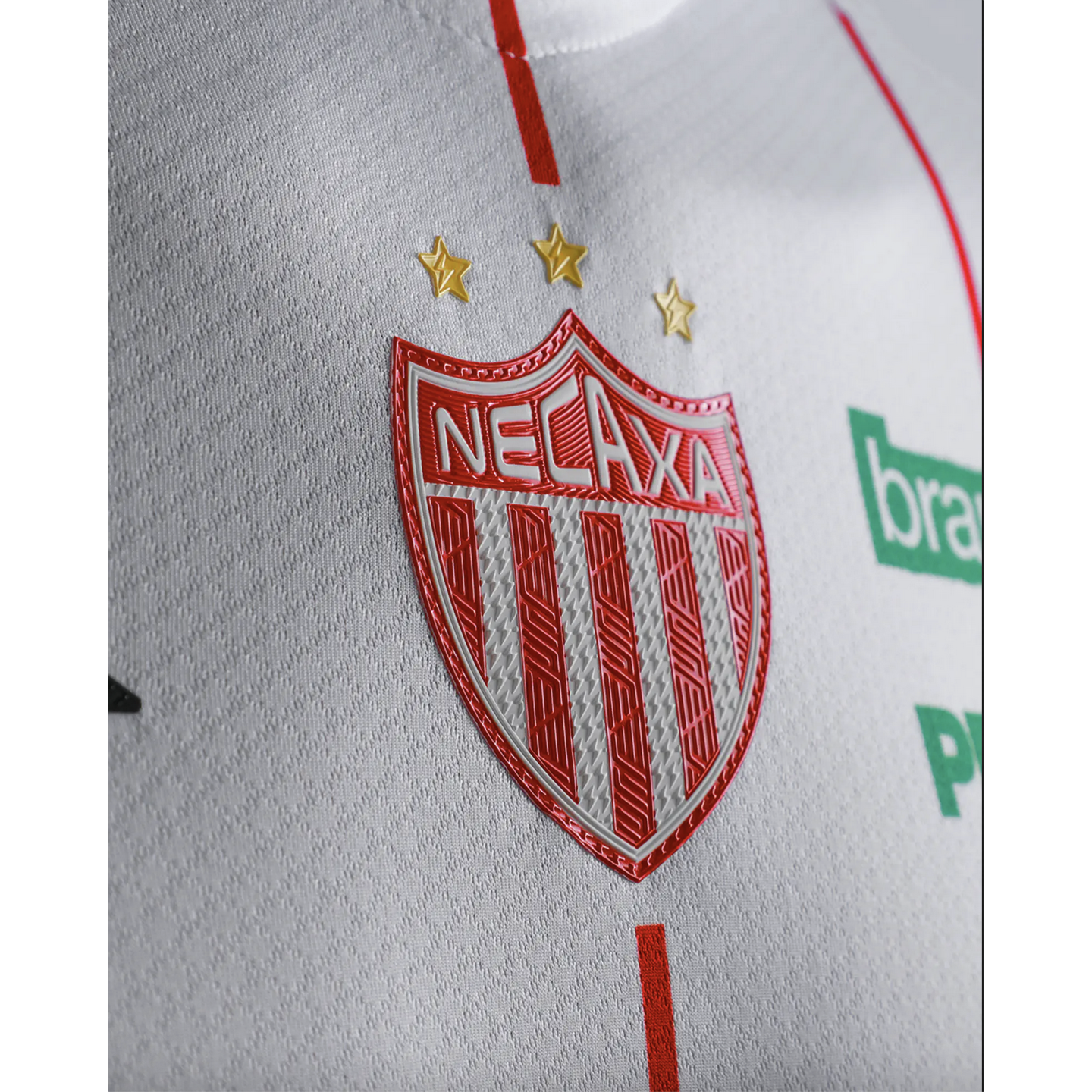 Close-up of a white textured Necaxa soccer jersey featuring a red and white shield-shaped team crest with vertical stripes and three gold stars above it.