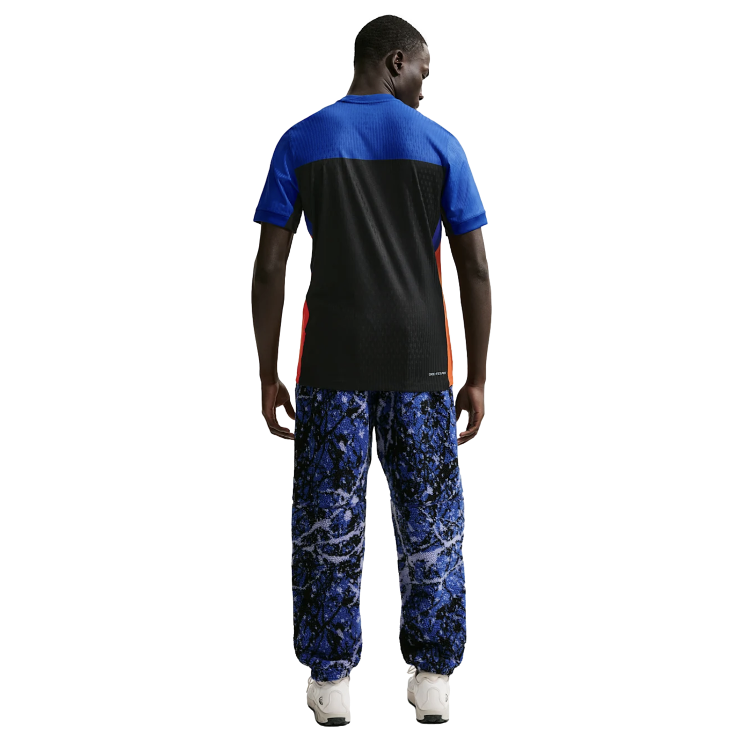 Back view of a man in a blue and black t-shirt and blue marbled print trousers with white sneakers, against a solid black background.