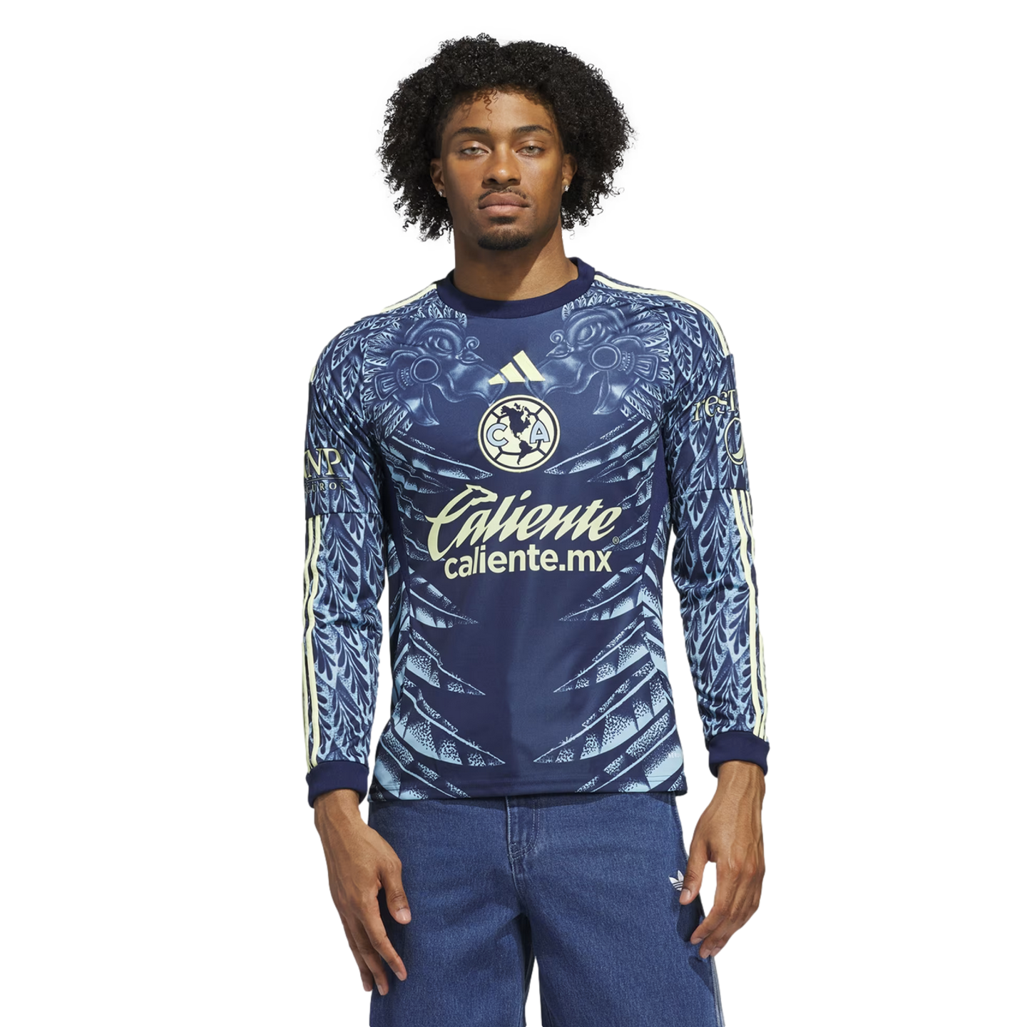 A man models a blue long-sleeved Club América soccer jersey with ornate light blue patterns and "Caliente" logo, wearing blue jeans against a black background.