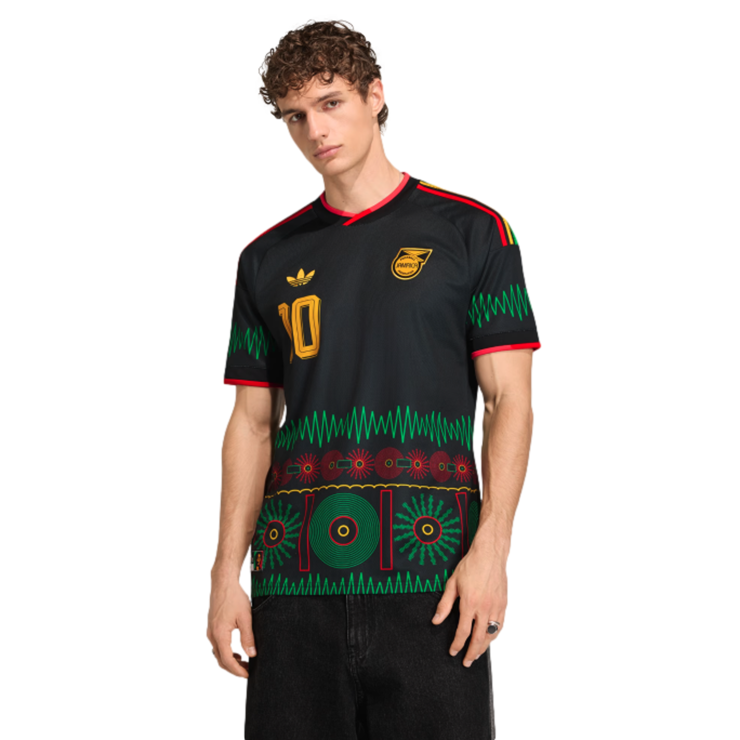 A man models a black Adidas Jamaica soccer jersey with green, red, and yellow patterns of vinyl records and sound waves.