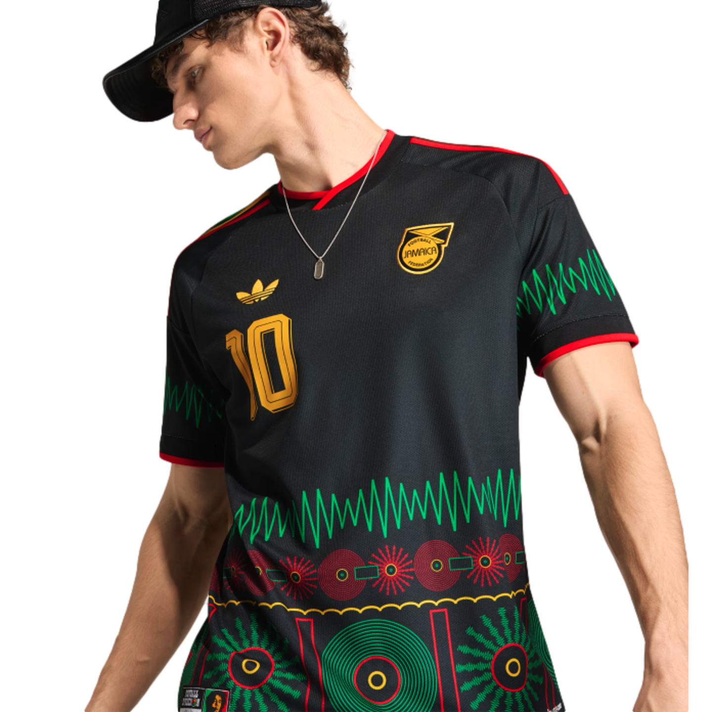 Man in a black Jamaica football jersey with vibrant green and red graphic patterns, a gold number 10, and team crest. He wears a black cap and silver necklace.