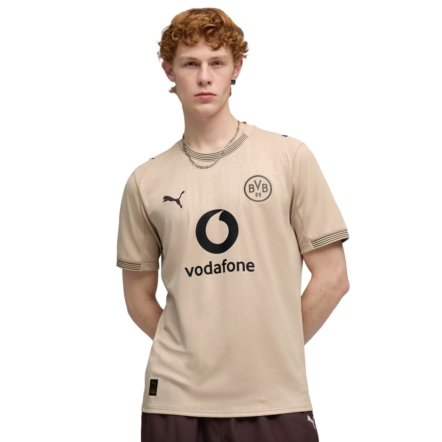 A man with curly red hair wearing a beige Borussia Dortmund soccer jersey with a Vodafone logo, posing against a black background.