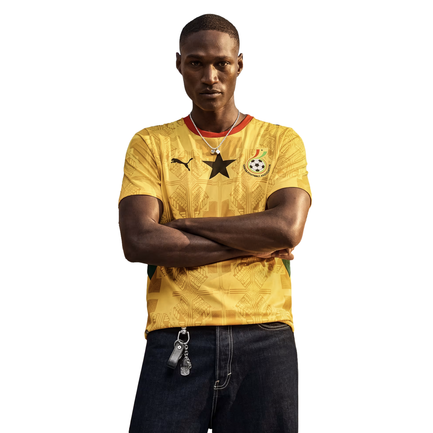 A man with his arms crossed stands against a black background, wearing a yellow Ghana national team soccer jersey and dark jeans.