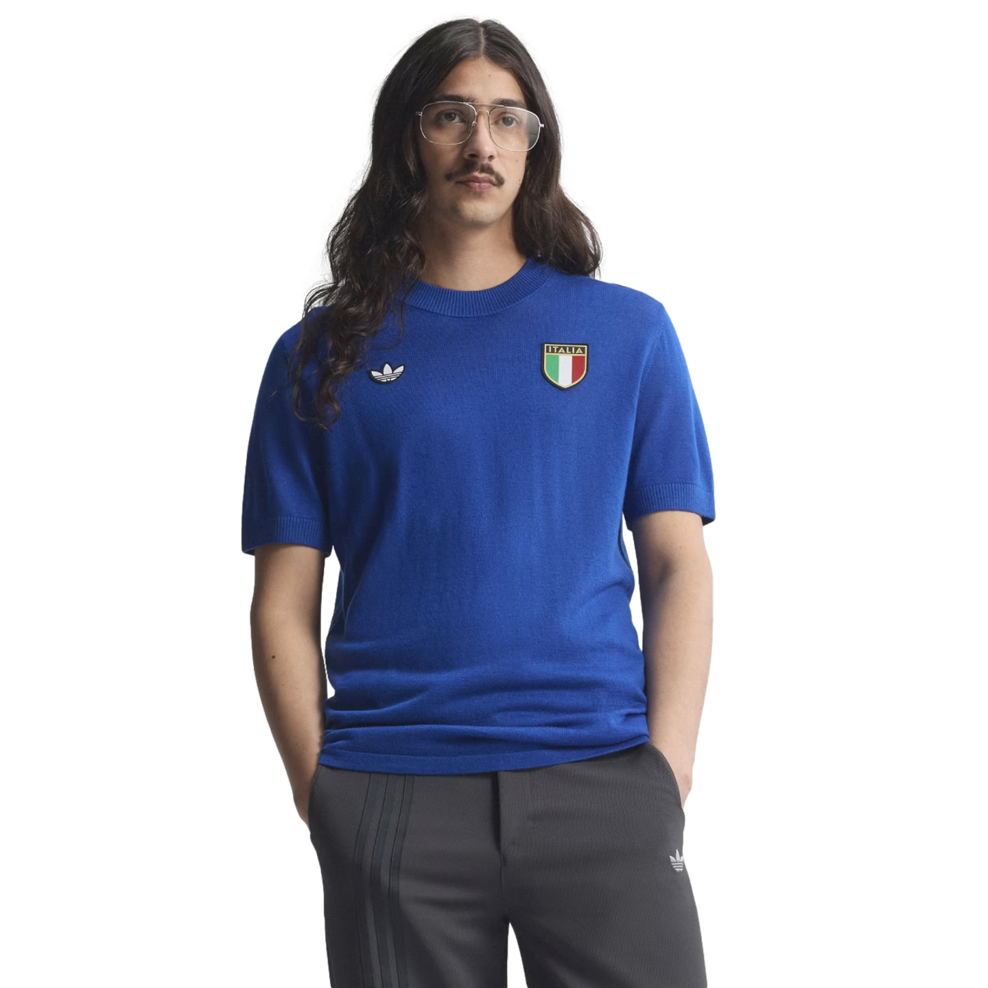 A man with long dark hair, glasses, and a mustache wearing a blue Adidas Italia knit shirt and grey trousers against a white background.