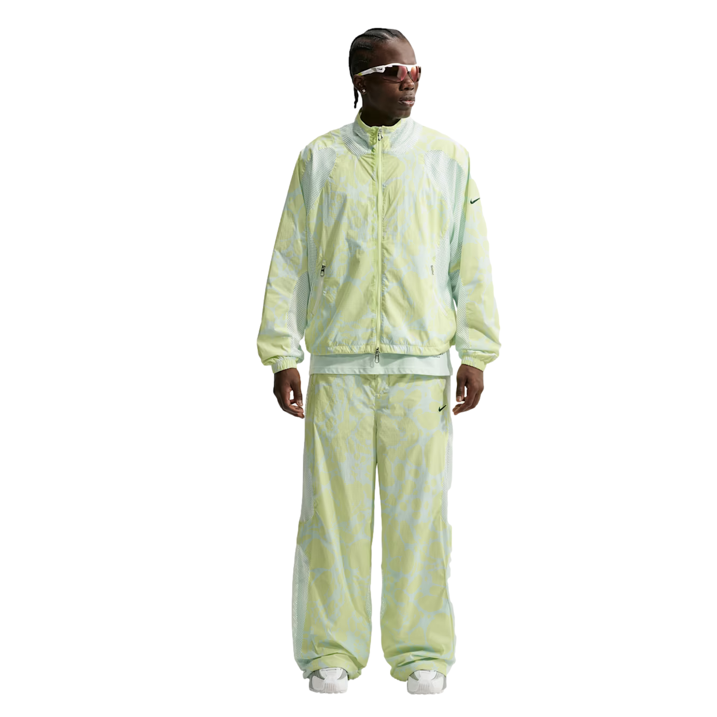 A person wearing a pale green patterned Nike tracksuit with wide-leg pants, white sunglasses, and white sneakers against a white background.