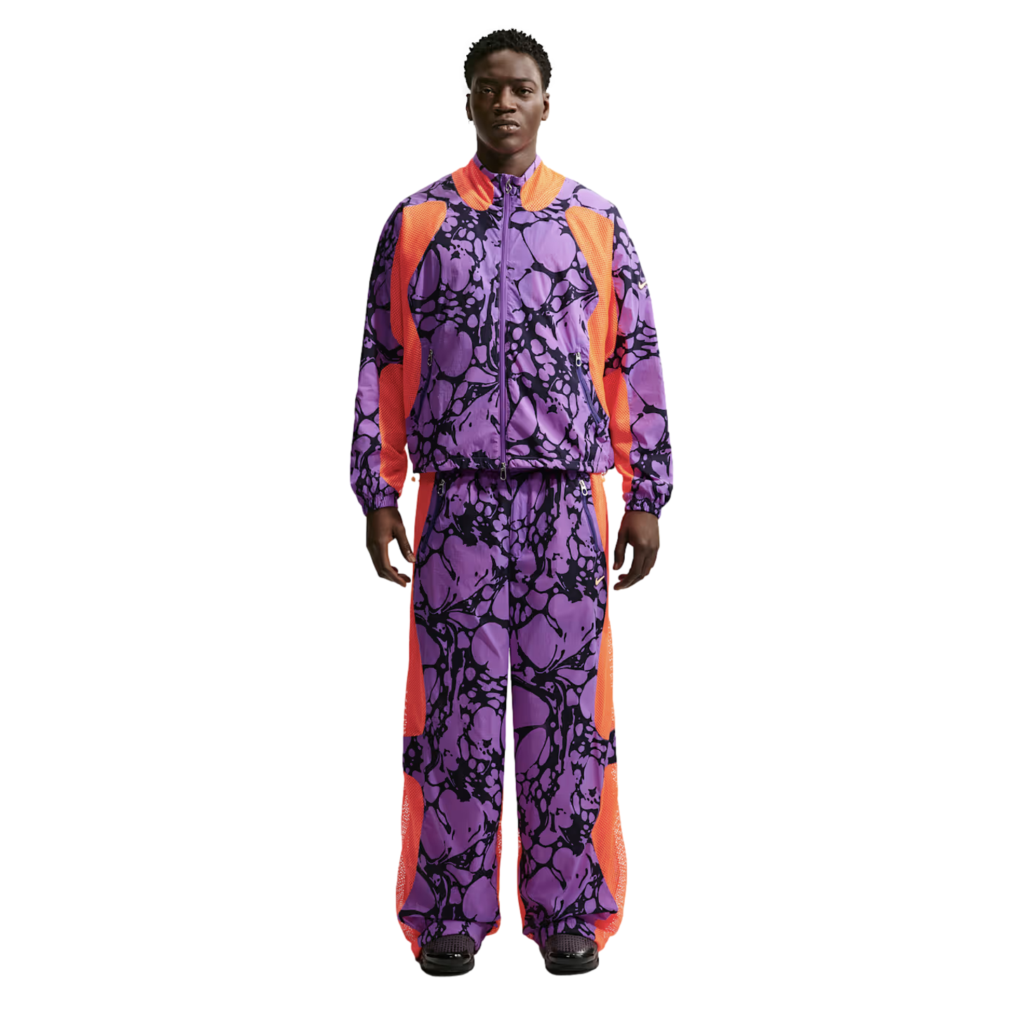 A Black man models a vibrant purple tracksuit with black marble patterns and bright orange mesh side panels, standing against a white background.