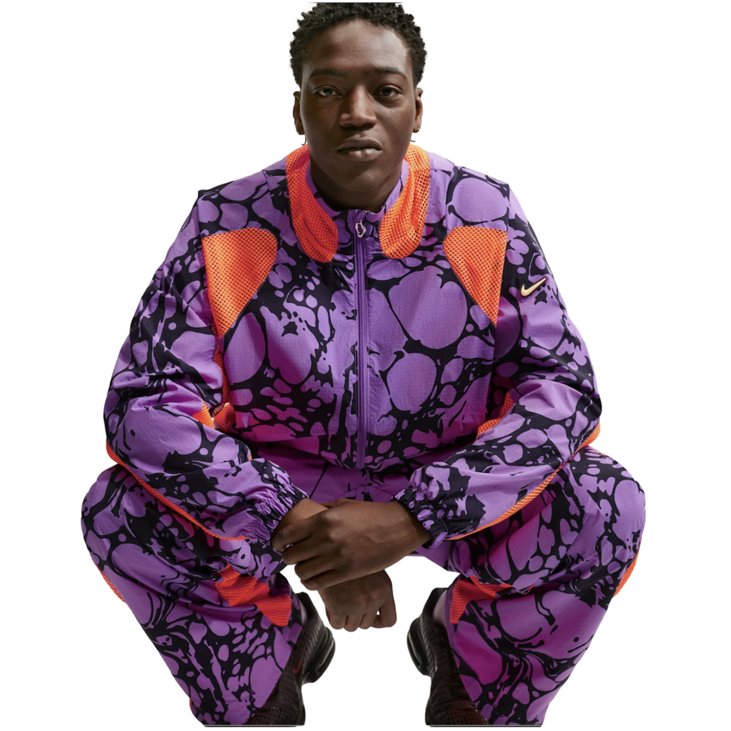 A person in a purple and black patterned Nike tracksuit with orange mesh panels, squatting against a white background.