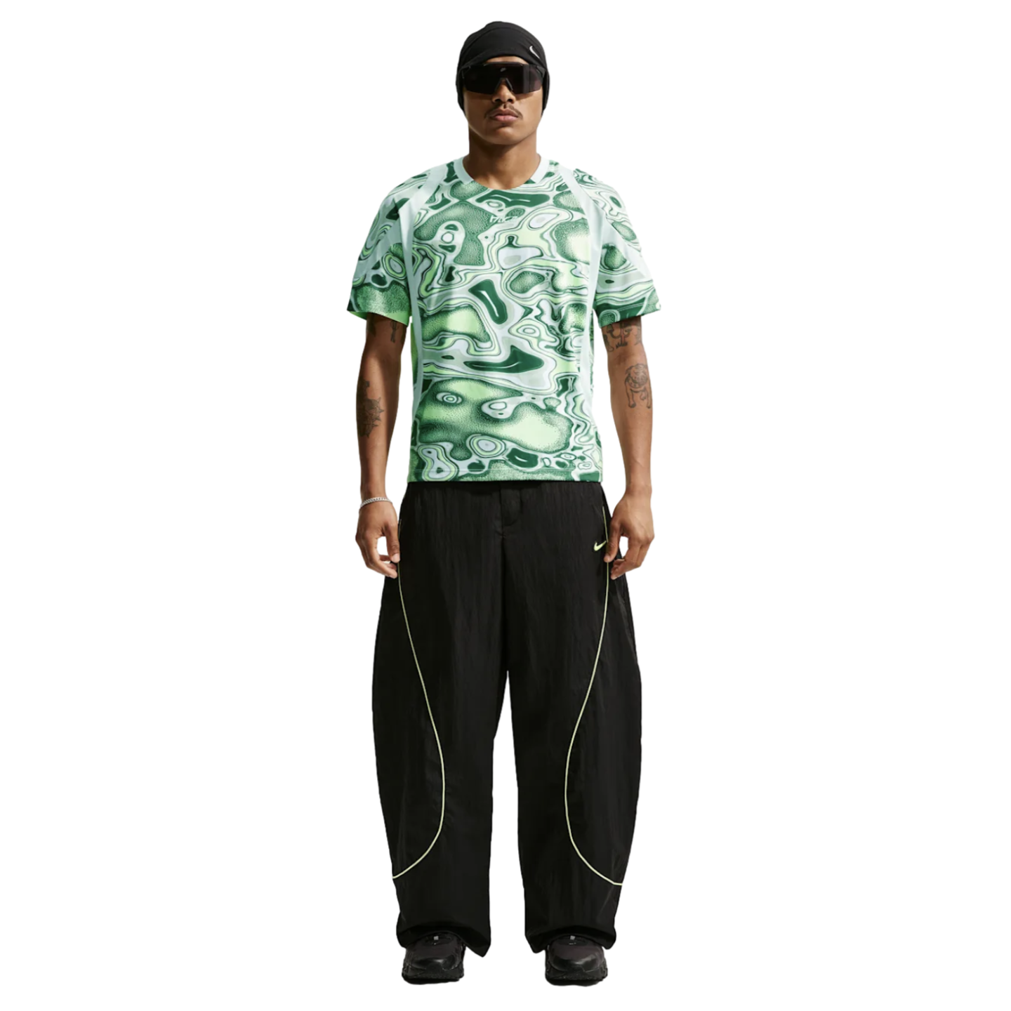 Man wearing a green patterned Nike shirt, baggy black pants with lime yellow piping, a black beanie, and sunglasses against a plain white background.