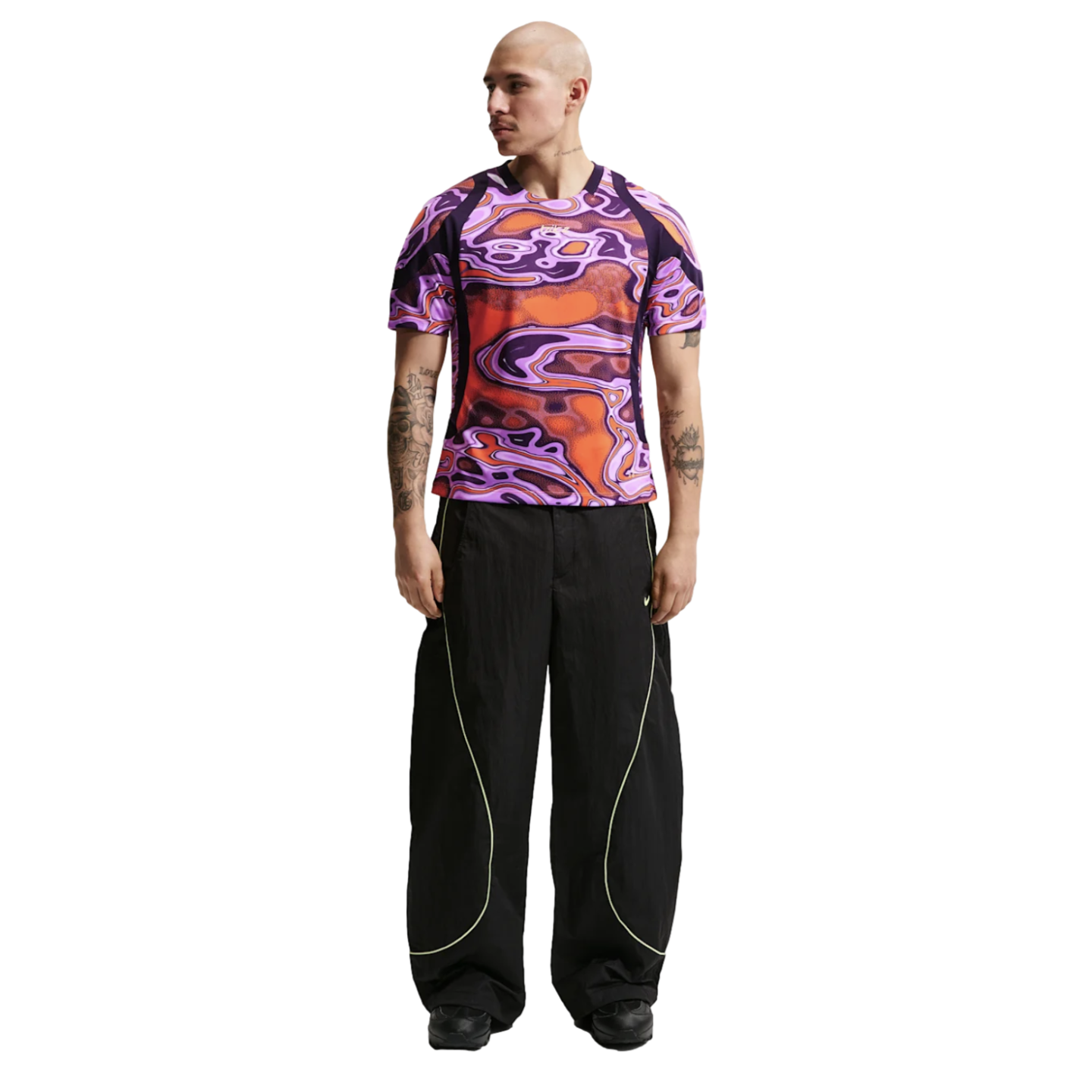 A man wearing a purple and orange psychedelic-patterned t-shirt and black wide-leg pants with neon piping, standing against a white background.