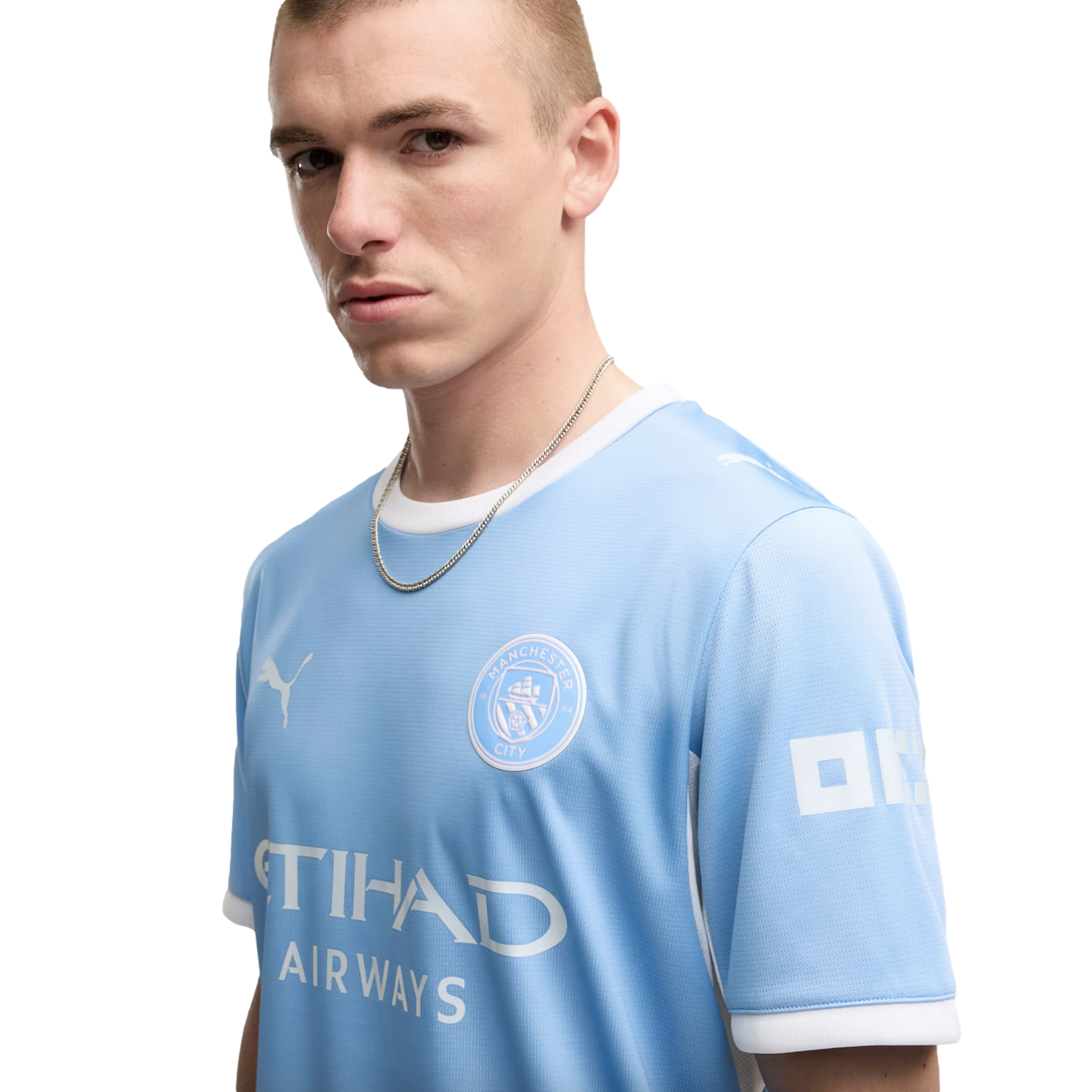 A young man with a short buzz cut wearing a light blue Manchester City soccer jersey and a silver chain poses against a white background.