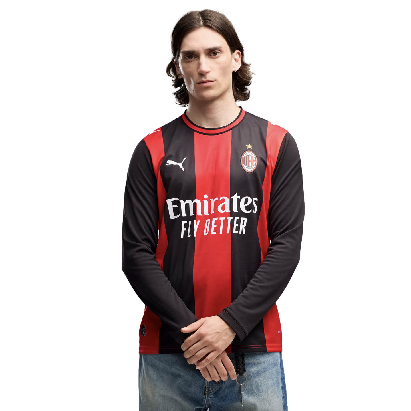 A man with mid-length dark hair wearing an AC Milan long-sleeved soccer jersey and blue jeans, standing against a plain white background.